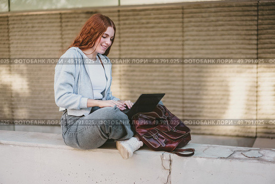 A young woman casually sits on a ledge, engrossed in her laptop. This scene perfectly embodies themes of productivity, remote work, and the contemporary lifestyles found in urban environments. A young woman working diligently on her laptop outdoors, dressed in stylish casual attire. Стоковое фото, агентство Ingram Publishing / Фотобанк Лори