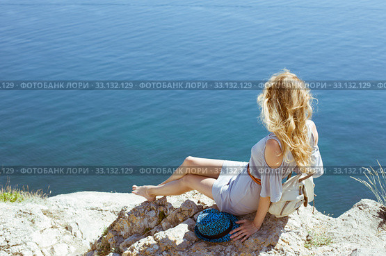 A young woman in a summer dress sits on the edge of a mountain or cliff with a beautiful sea view. Стоковое фото, фотограф katalinks / Фотобанк Лори