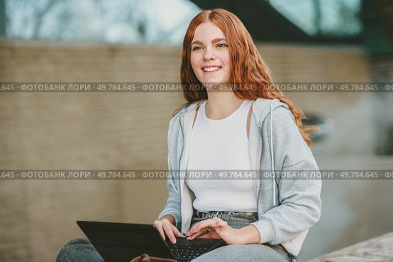 A young woman with long red hair sits outdoors with a smile, working on her laptop. She radiates positivity and productivity in a vibrant setting, reflecting her carefree yet focused lifestyle. A cheerful young woman happily working on her laptop outdoors in a vibrant, sunny setting. Стоковое фото, агентство Ingram Publishing / Фотобанк Лори