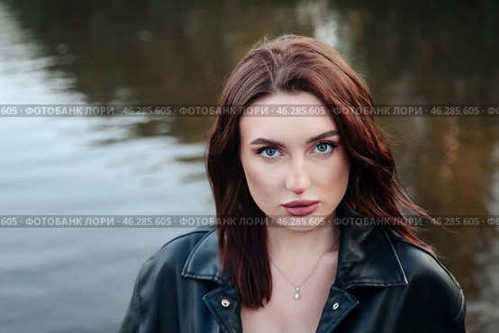 A young woman with striking blue eyes stands confidently by calm waters, wearing a dark leather jacket. The warm glow of sunset illuminates her features, creating a serene yet intense atmosphere. Стоковое фото, фотограф Aleksandr Golubev / Фотобанк Лори