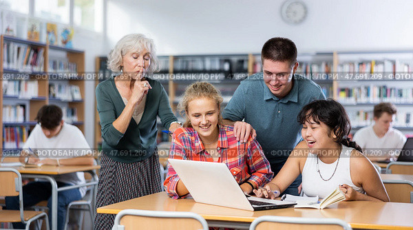 Adult librarian holding his finger on lips making silent gesture in library to laughing teenage pupils watching online video. Стоковое фото, фотограф Яков Филимонов / Фотобанк Лори