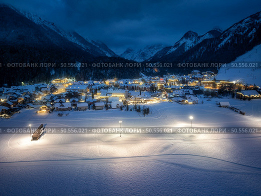 Aerial view of beautiful alpine village at cold winter night. Kranjska Gora, Slovenia. Top drone view of buildings in snow, street lights, road, snowy alpine mountains, sky at dusk. Triglav park. Стоковое фото, агентство Ingram Publishing / Фотобанк Лори