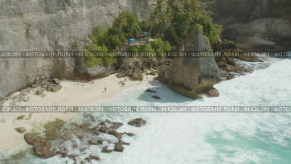 Aerial view of Diamond Beach in Nusa Penida, showcasing a small secluded sandy cove with tourists exploring near rocky cliffs. Стоковое видео, видеограф Ольга Сухарева / Фотобанк Лори