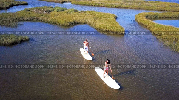 Aerial view of man and woman stand up paddleboarding on lake. Young couple are doing watersport on lake. Male and female tourists are in swimwear during summer vacation. Стоковое фото, агентство Ingram Publishing / Фотобанк Лори