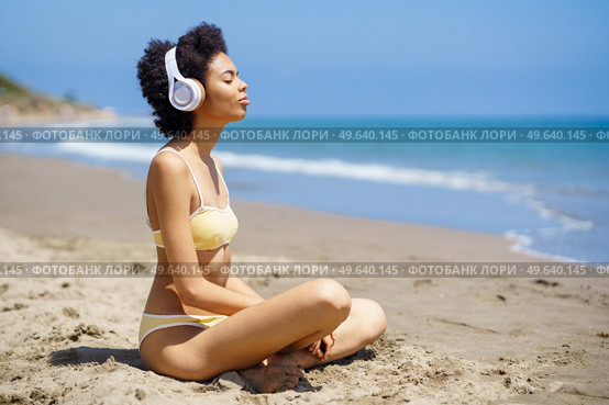African American woman sitting on the beach doing mindfulness listening to a meditation with wireless headphones.. Black woman sitting on the beach doing mindfulness listening to a meditation with headphones. Стоковое фото, агентство Ingram Publishing / Фотобанк Лори