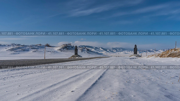 An asphalt road runs through a snow-covered valley. There are figures of idols on the roadsides. Ahead is a mountain range against the blue sky. Perspective. Altai. Chuysky tract. Стоковое фото, фотограф Вера Ивановна Кубарева / Фотобанк Лори