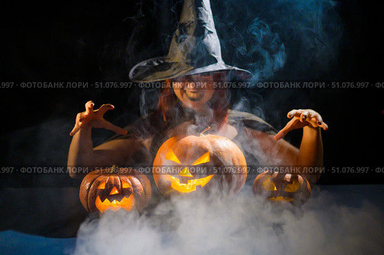 An ominous witch in a hat conjures over a jack-o-lantern. Traditional halloween characters. Mystical fog creeps over pumpkins with carved terrible faces. Woman in carnival costume. Стоковое фото, фотограф Михаил Решетников / Фотобанк Лори