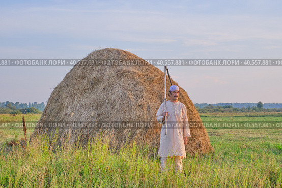 An senior Muslim man in a skullcap and traditional clothes leaves with a hand scythe from the hay field at sunset. Стоковое фото, фотограф Ирина Кононова / Фотобанк Лори