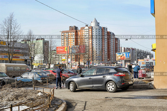 An urban street landscape on a clear sunny day. In the foreground is a parked dark gray hatchback and several people, one of whom is standing next to the car, and the other is walking towards the parking lot. On the left are rows of parked cars and a section of fencing separating the pedestrian zone from the roadway. In the background are modern multi-story residential buildings with red brick and gray metal facades, as well as older panel houses in green and gray. Billboards and store signs, including a supermarket and an optician, are installed along the street. On the sidewalk on the right, several people are busy with their own affairs. In the foreground, the remains of dirty snow are noticeable, typical for late winter or early spring. (2013 год). Редакционное фото, фотограф Анна Кривицкая / Фотобанк Лори