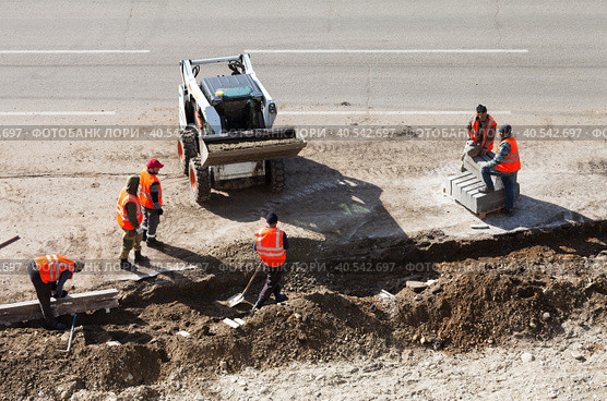 ANGARSK, IRKUTSK REGION, RUSSIA - May 18, 2023: Workers in signal red vests lay a curb stone on the side of a road being repaired. Mini tractor loader brings soil in the bucket. Редакционное фото, фотограф Виктория Катьянова / Фотобанк Лори