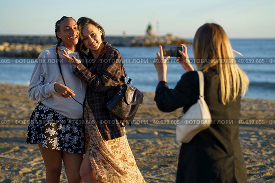 Anonymous woman taking photo of positive multiracial female friends hugging while standing on sandy coast near rippling sea on sunny day. Faceless woman photographing cheerful diverse girlfriends on seashore. Стоковое фото, агентство Ingram Publishing / Фотобанк Лори