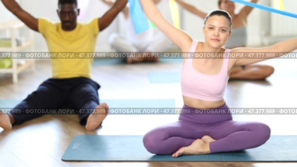 Asian girl conducts Pilates multinational group sitting, elastic band in hands above head body bends. Стоковое видео, видеограф Яков Филимонов / Фотобанк Лори