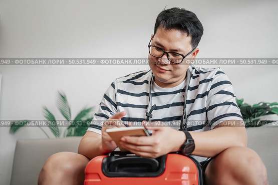 Asian man glasses is preparing clothes in suitcases. He’s choosing clothes, travel documents, the itinerary for a solo trip, and check the checklist. Travel, holiday and vacation concept. Стоковое фото, агентство Ingram Publishing / Фотобанк Лори
