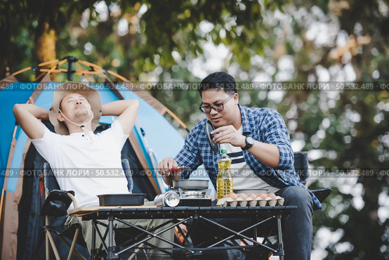 Asian relaxed man using smartphone and waiting for his friend cooking with tent background in camping. Cooking set front ground. Outdoor cooking, traveling, camping, lifestyle concept. Стоковое фото, агентство Ingram Publishing / Фотобанк Лори