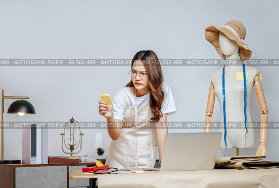 Asian young pretty female fashion designer stylish wearing eyeglasses standing and serious with paper not during working at fashion studio, full of tailoring tools and equipment. Стоковое фото, агентство Ingram Publishing / Фотобанк Лори