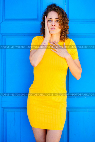 Astonished young female in yellow dress with curly brown hair keeping hand on chest and touching face while looking away against blue wall on street. Amazed woman touching chest and face on street. Стоковое фото, агентство Ingram Publishing / Фотобанк Лори