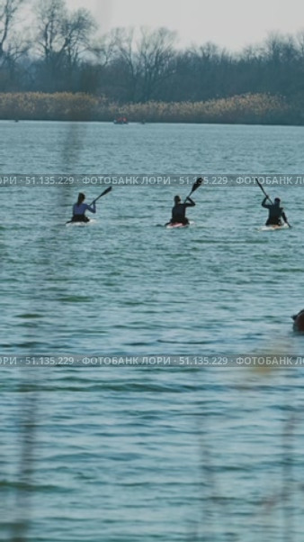 Athletes kayak on a calm lake framed by distant trees and reeds. Soft ripples reflect the clear sky. Preparation of athletes a training base. View from behind. Стоковое видео, видеограф Воловодов Владимир / Фотобанк Лори