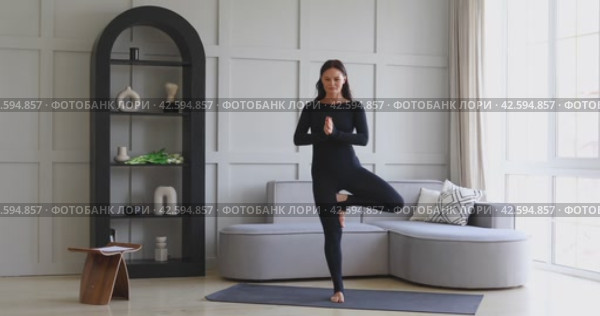 Attractive athletic woman performs various variations of Vrikshasana exercise at home, tree pose, training in black overalls while standing on the mat, full length view, yoga concept. Стоковое видео, видеограф Сергей Чайко / Фотобанк Лори