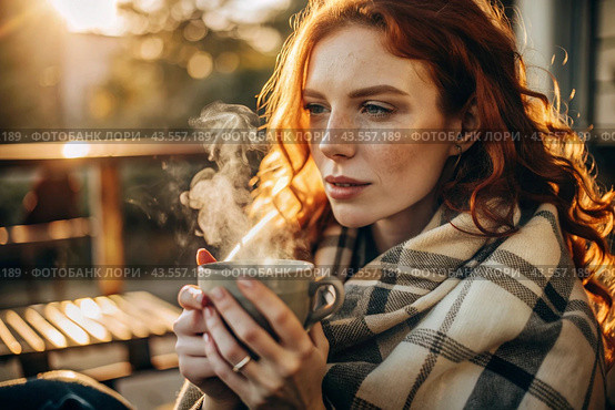 Autumn evening. A red-haired, beautiful woman wrapped in a plaid with a mug of hot coffee on a beautiful patio in the rays of the setting sun. The girl inhales the aroma of the drink. Romance, comfort and harmony. Стоковое AI-изображение, AI-художник Photojuli / Фотобанк Лори