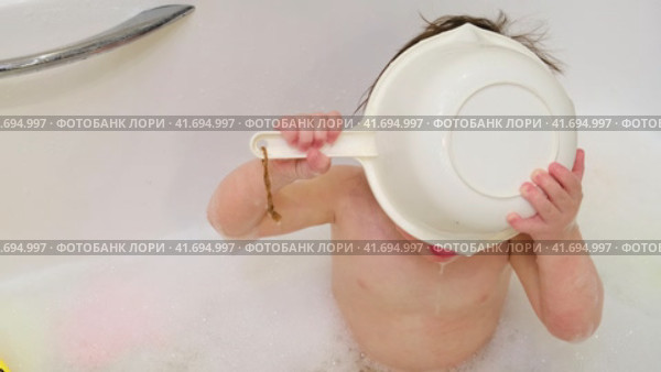 Baby drinks water from a ladle while sitting with toys in a white bathtub A happy child enjoys washing in the bathroom. Kid about two years old (one year ten months). Стоковое видео, видеограф Андрей Журавлев / Фотобанк Лори