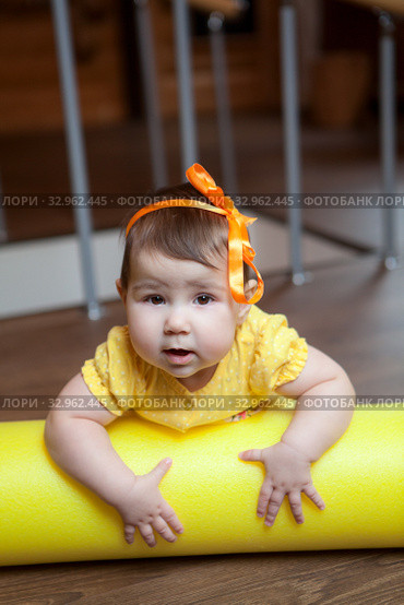 Baby girl lying on yellow foam roller in domestic gym, looking at camera. Стоковое фото, фотограф Кекяляйнен Андрей / Фотобанк Лори