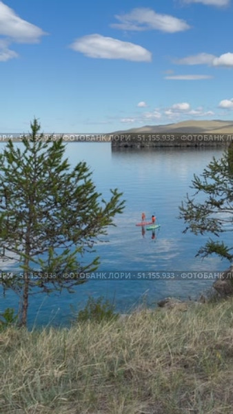 Baikal Lake in summer. Vertical video of two girls in orange life jackets and hats riding SUP boards on blue water of Small Sea on sunny day. Active seaside recreation. Стоковое видео, видеограф Виктория Катьянова / Фотобанк Лори