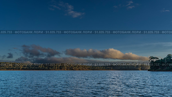 Beautiful calm lake. Ripples on the blue water. Green vegetation on the shores. The clouds in the sky are illuminated by the golden evening light. Madagascar. Lake Mantasoa. Стоковое фото, фотограф Вера Ивановна Кубарева / Фотобанк Лори