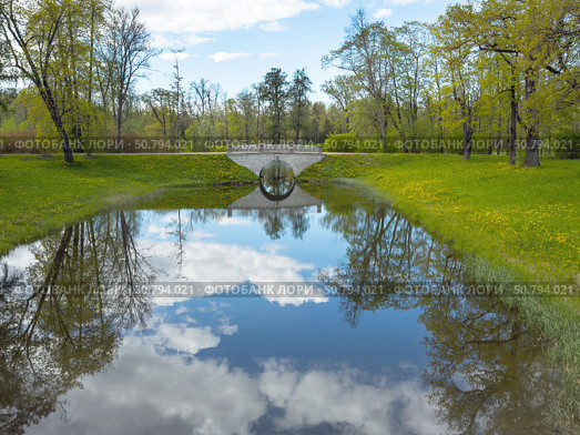 Beautiful deserted park in spring. View of stone pedestrian bridge with mirror reflection in quiet river. Green beds with dandelions and fresh young foliage on trees. Picturesque landscape of sunny May day. Стоковое фото, фотограф Виктория Катьянова / Фотобанк Лори