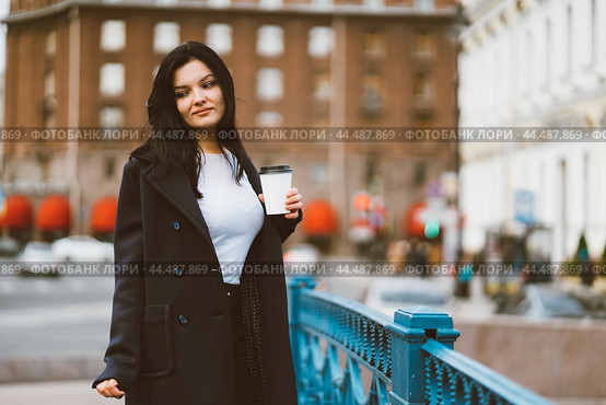 Beautiful serious smart brunette girl holding cup of coffee in hands goes walking down street in center on blue bridge. Charming thoughtful woman with long hair wanders alone, immersed in thoughts. Стоковое фото, фотограф Natalia Gdovskaia / Фотобанк Лори