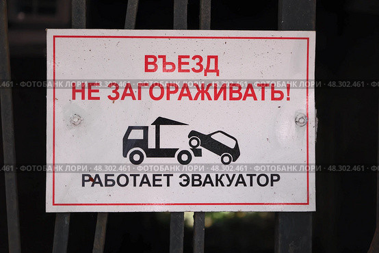 Белый прямоугольный знак с красной каймой и надписью: "ВЪЕЗД НЕ ЗАГОРАЖИВАТЬ! РАБОТАЕТ ЭВАКУАТОР" и иконкой эвакуатора. На темных металлических воротах. Стоковое фото, фотограф Евгений Ковальский / Фотобанк Лори