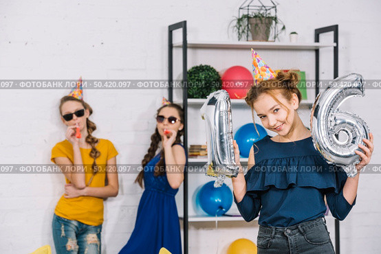 birthday girl showing numeral 16 foil silver balloons with her two friends blowing party blower. Стоковое фото, агентство Ingram Publishing / Фотобанк Лори