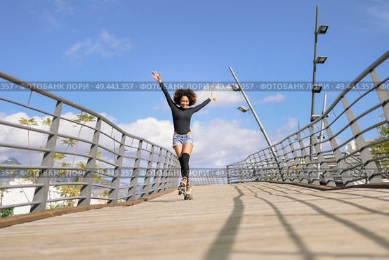 Black woman, afro hairstyle, on roller skates riding outdoors on urban bridge with open arms. Smiling young female rollerblading on sunny day. Beautiful clouds in the sky.. Afro hairstyle woman on roller skates riding outdoors on urban bridge. Стоковое фото, агентство Ingram Publishing / Фотобанк Лори