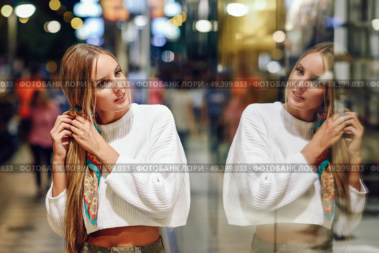 Blonde girl wearing white sweater standing in the street with defocused city lights at the background. Pretty woman with pigtail hairstyle at night reflected in a shop window. Стоковое фото, агентство Ingram Publishing / Фотобанк Лори