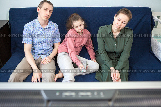 Boring Caucasian family sitting on couch spending time together by watching television set, people staying at home. Стоковое фото, фотограф Кекяляйнен Андрей / Фотобанк Лори