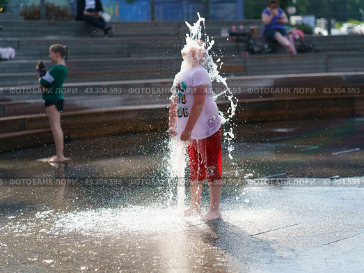 Boy washing hif face by cold water of fountain in the hot summer day in the big city. Fresh water splashes. He is wet. Concept of the leisure time, freshness and happiness. Frontal view (2021) Редакционное фото, фотограф YuryKara / Фотобанк Лори