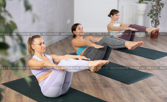 Calm sportive women performing yoga boat pose on mats Paripurna Navasana during group training in fitness studio. Стоковое фото, фотограф Яков Филимонов / Фотобанк Лори