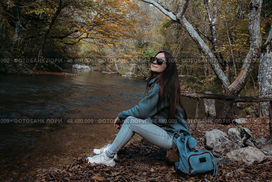 Caucasian girl with long dark hair sits on the river bank. Hiking. Стоковое фото, фотограф София Тюленева / Фотобанк Лори