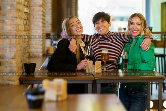 Cheerful adult female with short dark hair looking at camera with smile and embracing girlfriends behind table with alcohol during meeting in pub. Happy women hugging during party in bar. Стоковое фото, агентство Ingram Publishing / Фотобанк Лори