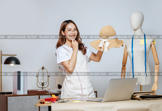 Cheerful Asian young female designer wearing eyeglasses doing fashion design on beautiful sombrero in studio, full of tailoring tools  with mannequin and equipment on the desk. Стоковое фото, агентство Ingram Publishing / Фотобанк Лори