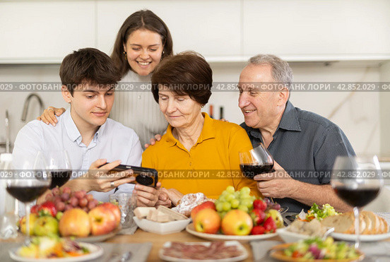 Cheerful family browsing photos on smartphone at home dinner. Стоковое фото, фотограф Яков Филимонов / Фотобанк Лори