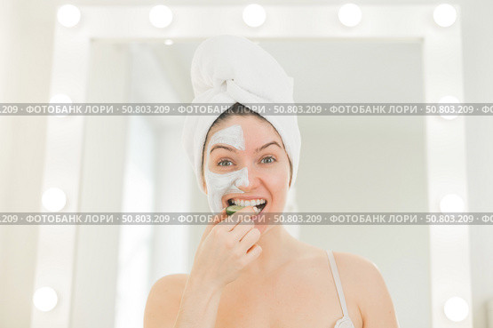 Cheerful woman with a towel on her hair and in a clay face mask fooling around with cucumbers in her hands. Taking care of beauty at home. Стоковое фото, фотограф Михаил Решетников / Фотобанк Лори