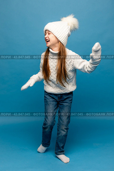 Child girl in a white sweatshirt and knitted hat with pompom on a blue background. The concept of children's emotions, joy and delight. Advertising clothing and sales. Ski resort vacation, copy space. Стоковое фото, фотограф Ирина Михайличенко / Фотобанк Лори