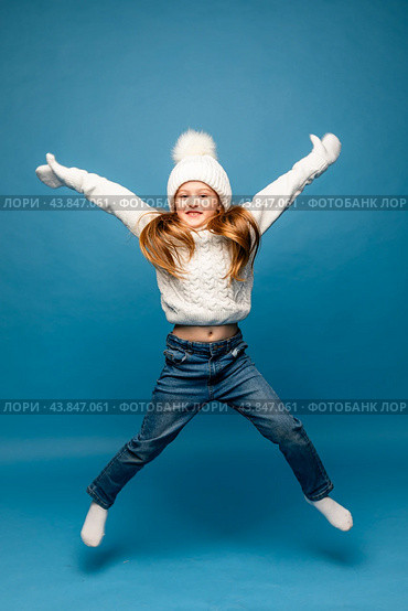 Child girl in a white sweatshirt and knitted hat with pompom jumping on a blue background. The concept of children's emotions, joy and delight. Advertising clothing and sales. Ski resort vacation. Стоковое фото, фотограф Ирина Михайличенко / Фотобанк Лори