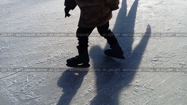Children ride in a city park on an ice rink. Feet skater while skating on ice. The low winter sun weakly illuminates the ice. Dark shapes and long shadows on the surface. Sports movements. Стоковое фото, фотограф Юлия Петровская / Фотобанк Лори