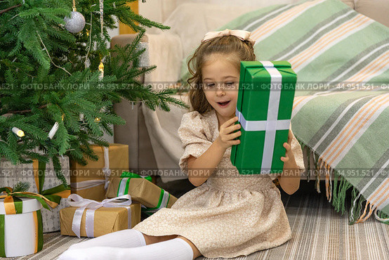 Christmas spirit. Happy little smiling girl opening gift box sitting on the floor near the Christmas tree. Happy girl unwrapping Christmas present looking in box. Holidays and celebrations concept. Стоковое фото, фотограф Ирина Михайличенко / Фотобанк Лори
