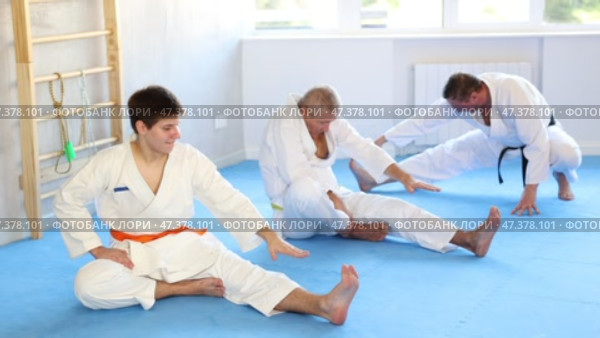 Concentrated adolescent jiu-jitsu fighter doing stretching exercises on mat, warming up body muscles before training during group martial arts class. Стоковое видео, видеограф Яков Филимонов / Фотобанк Лори