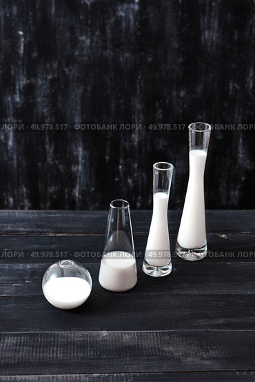 Containers of glass cone and spherical shape, filled with white liquid on a dark wooden table on a black background with copy space. Milk saving technology.. Diagonal composition of glass of different shapes and sizes filled with milk or yogurt on a black background. Natural organic dairy product. Стоковое фото, агентство Ingram Publishing / Фотобанк Лори