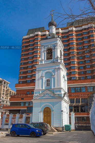 Contrast between the historical and modern architecture of Moscow. In the foreground is the bell tower of an ancient church, painted in blue and white, topped with an onion-shaped dome with a cross. Church bells hang in the opening of the tower.In the background rises the President Hotel, a monumental Soviet building with its characteristic beige brick cladding, many windows and cascading structure.This image perfectly demonstrates the coexistence of religious heritage and Soviet modernism, typical of Moscow. Стоковое фото, фотограф Анна Кривицкая / Фотобанк Лори