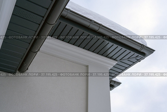 Corner of the house with new gray metal tile roof and rain gutter covered by snow at winter. Metallic Guttering System, Guttering and Drainage Pipe Exterior. Стоковое фото, фотограф Данила Большаков / Фотобанк Лори
