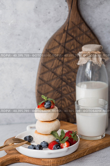 Cottage cheese pancakes, Russian cheesecakes with fresh blueberries, strawberries and mint on a stylish wooden board on a gray concrete table. Natural products. Healthy and delicious breakfast. Стоковое фото, фотограф Photojuli / Фотобанк Лори